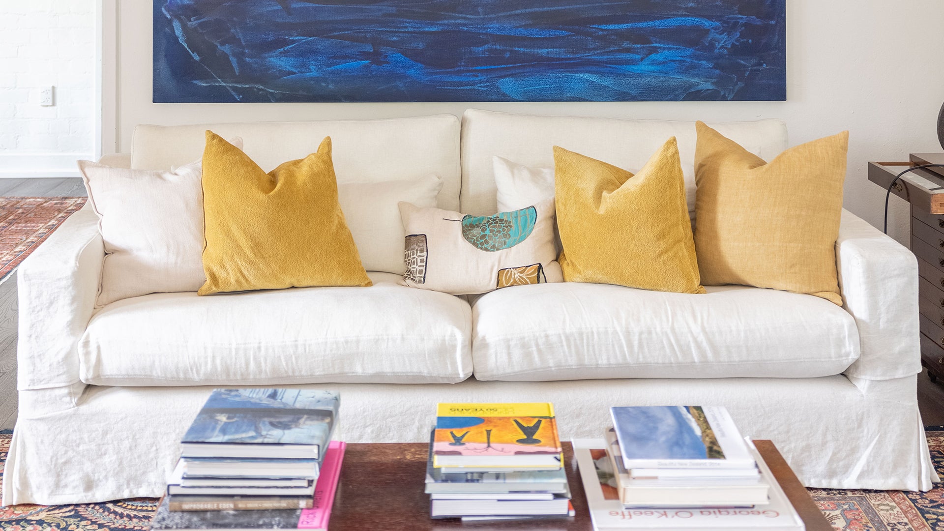 A white sofa with mustard cushions with a dark blue piece of artwork behind it, and a wooden coffee table in-front with a bunch of books sitting on top.