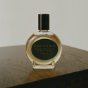 A round glass perfume bottle filled with amber liquid, featuring a black cap and a dark green label with gold lettering that reads 'CURIONOIR' and 'OPIA'. The bottle is placed on a dark wooden surface against a light gray background.
