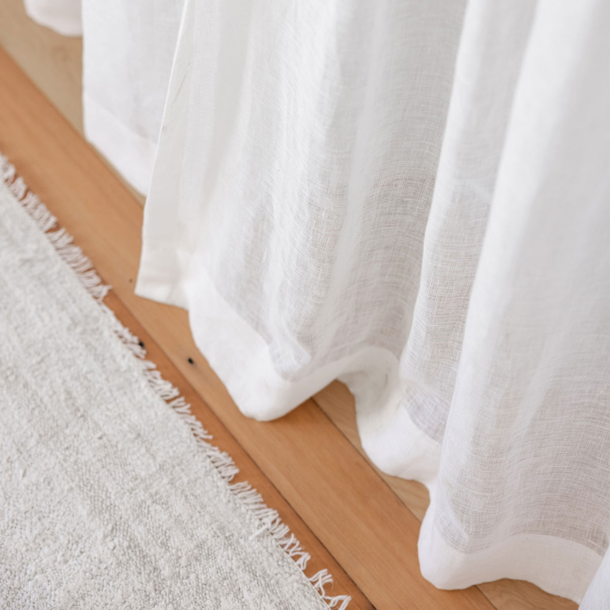 Looking at the bottom of a white sheer curtain on a wooden floor with a natural rug.