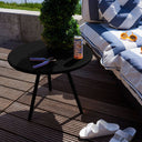 Black side table with a can of beer on a wooden deck, next to a striped outdoor sofa and white slippers.