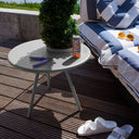 Outdoor setting with a small round grey table, plant, can, and sunglasses on a wooden deck.