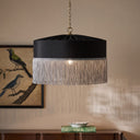 A black fabric drum pendant light with a silver fringe trim and a brass hanging chain, shown hanging in a styled room with a framed bird painting and a wooden console table.