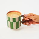 Hand holding a green and white checkered mug with a creamy beverage on a light background