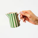 Hand holding a green and white striped ceramic pitcher against a light background