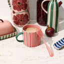 A striped ceramic mug with striped design, floral pitcher, and striped container on a light wooden surface.