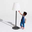 Child in blue overalls standing next to a tall lamp with a white shade and black base on a white background