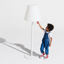 Child in blue overalls reaching up to a large white lampshade on a white background