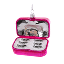 A novelty Christmas ornament shaped like an open pink lipstick case, containing three pairs of black false eyelashes on a white insert, with a silver hanging loop at the top.