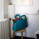 Teal storage bag with handles filled with vegetables on a wooden stool against a white wall.