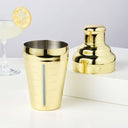 Gold cocktail shaker and bottle on a white surface with a glass of lemonade in the background.
