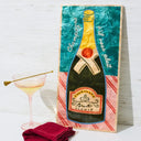 Decorative plaque with a champagne bottle design on a white surface with a cocktail and napkin.