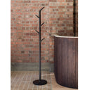 A black metal coat rack with a branch-like design stands on a tiled floor in front of a red brick wall, next to a dark wood barrel-style tub or bench.