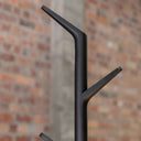 A close-up of a matte black metal coat rack with a tree-like branch design, set against a blurred brick wall background.