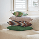 Four decorative throw pillows stacked on a light beige sofa in a bright room. The stack includes a green striped pillow on top, a plain beige pillow, a brown striped pillow, and a solid green pillow at the bottom.