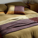 A cozy bedroom scene showing a bed made with olive green bedding, a matching pillow, and a folded throw blanket in earthy patchwork tones of mustard, rust, burgundy, and cream. A wooden bedside table and lamp are visible in the background.