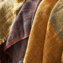 A close-up of a woven textile with draped folds, showing a color-blocked pattern in mustard yellow, deep burgundy, muted blue-gray, and tan with frayed edges.