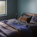 A styled bed in a bedroom with slate blue walls, featuring a dark gray linen bedding set with a blue and brown throw blanket, a brown pillow, and matching pillows.