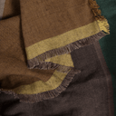 A close-up of a woven scarf or shawl with fringed edges, showing a color-blocked pattern in warm brown, olive yellow, and dark brown.