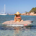 Woman on thew grey taupe Original Floatzac beanbag in the water, holding a watermelon slice with a sailboat and rocky shoreline in the background.