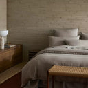 Bedroom with wooden dresser, bed with taupe duvet, and woven bench against a brick wall.