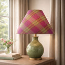Table lamp with a plaid pink and green shade on a wooden surface near a window.