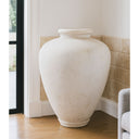 Large white textured vase on a wooden floor against a wall with a window.
